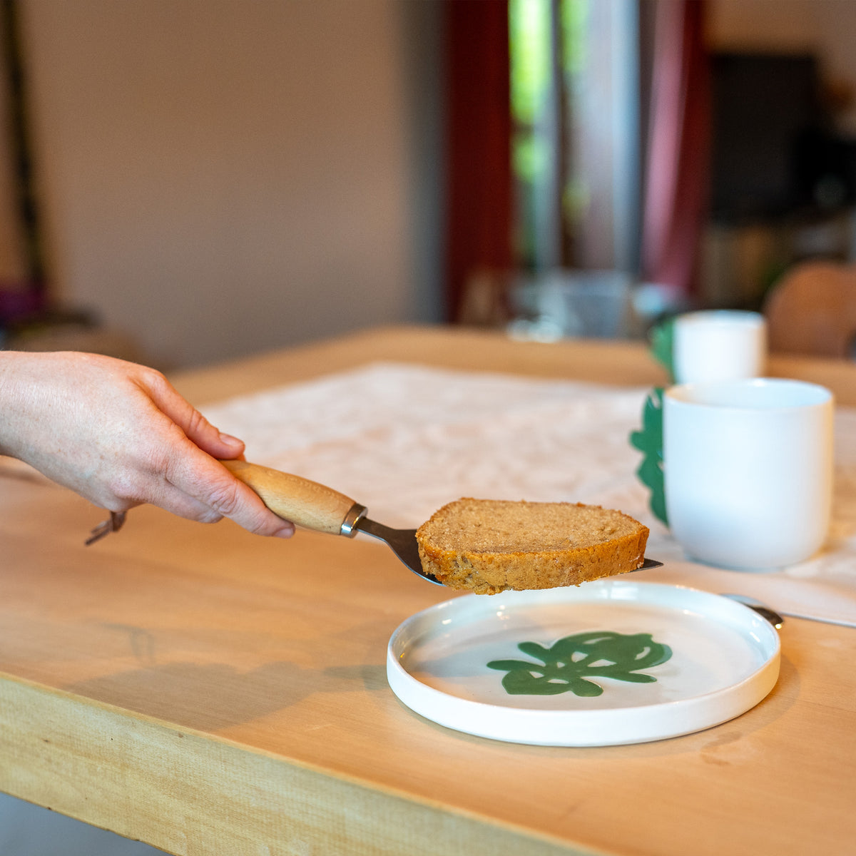 Assiette à dessert artisanale en porcelaine, fabriquée en France, blanche avec un motif cœur alsacien vert, sur table en bois avec tasses en porcelaine made in France