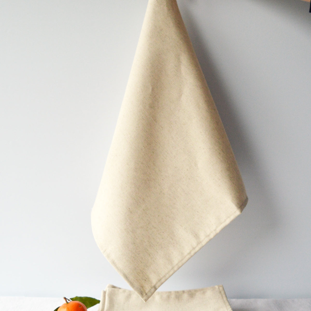 Serviette beige naturel en coton lin fabriquée en France suspendue au-dessus d'une pile de serviettes beiges pliées