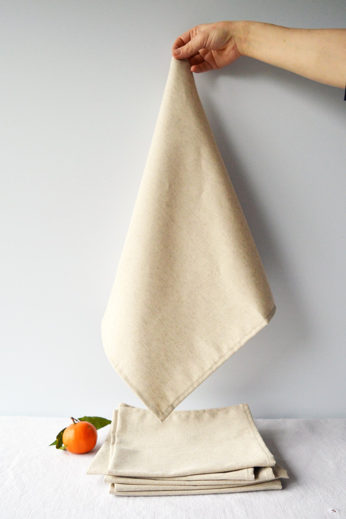 Serviette beige naturel en coton lin fabriquée en France suspendue au-dessus d'une pile de serviettes beiges pliées