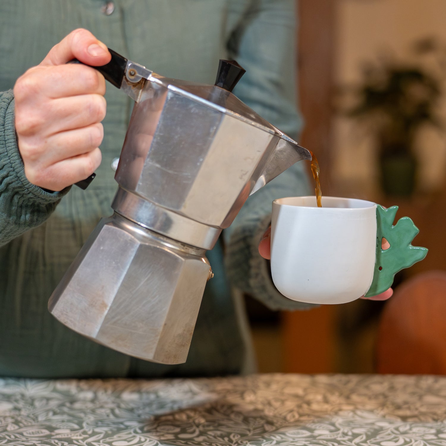 Tasse à café artisanale fabriquée en France, en porcelaine blanche avec une anse verte motif cœur alsacien, une cafetière italienne y verse du café