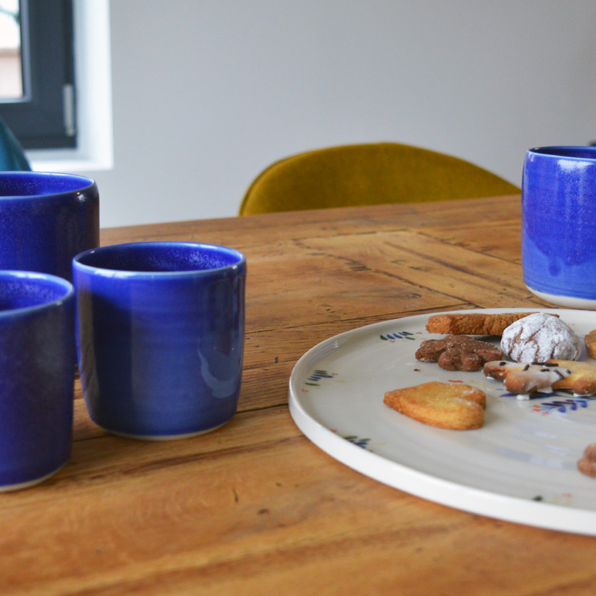 Porcelaine artisanale fabriquée en France, tasses à thé et à café bleues Maison Primavera, à côté d'un plat à motif Primavera, sur une table en bois.